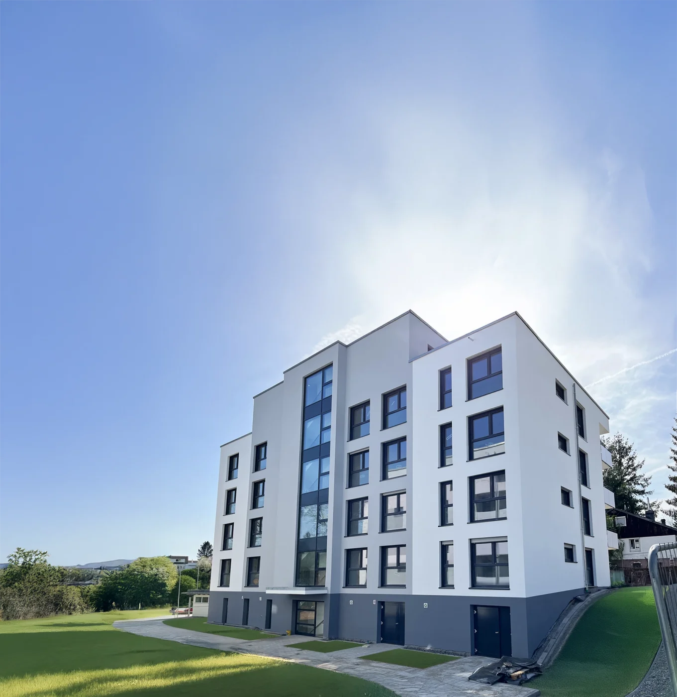 Bronnwiesenweg Rudersberg Ein modernes, weißes Neubau-Wohnhaus mit großen Fenstern hebt sich vor einem strahlend blauen Himmel ab, umgeben von grünem Rasen und Bäumen – eine ideale Immobilie für alle, die eine Immobilie kaufen möchten.