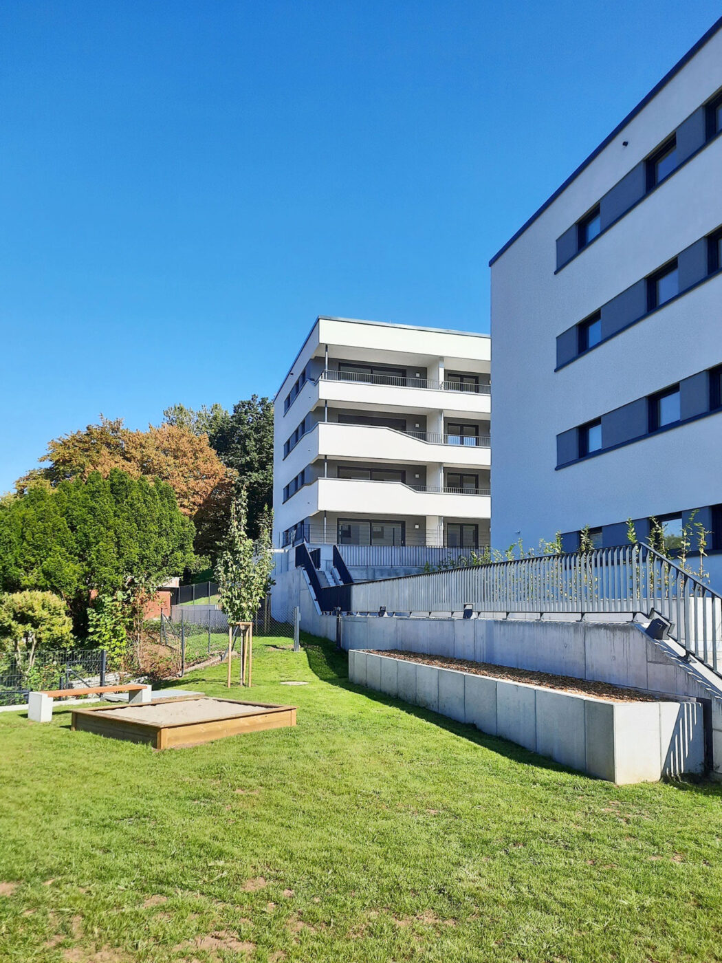 Schiesstal Schwäbisch Gmünd Moderne weiße Mehrfamilienhäuser mit Balkonen, umgeben von grünem Rasen, jungen Bäumen, einem Hochbeet und einem blauen Himmel über den Köpfen – ideal für alle, die eine Wohnung in Schorndorf kaufen oder eine Immobilie in einer lebendigen und zugleich ruhigen Umgebung suchen.
