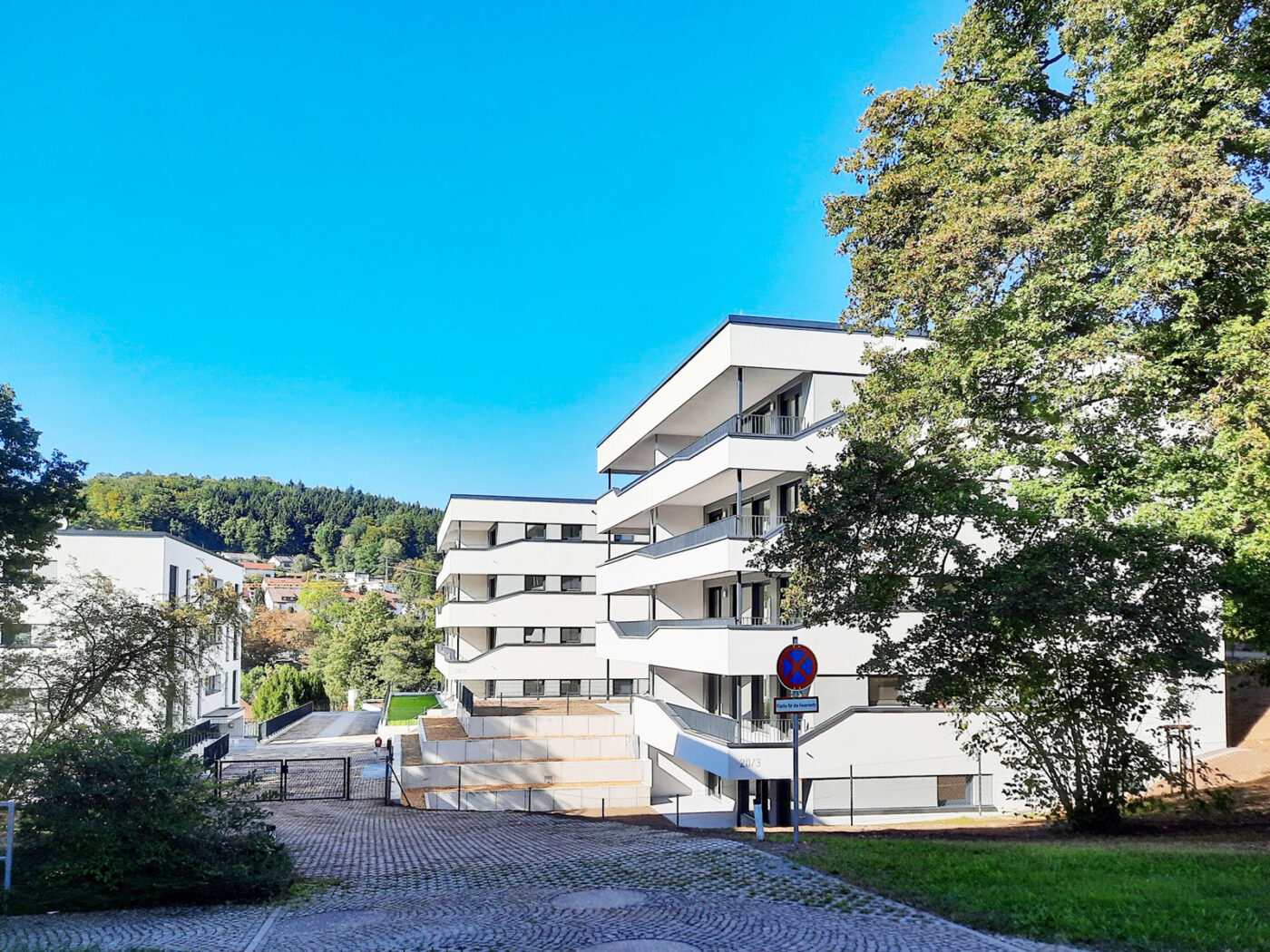 Energieeffiziente Neubauwohnung kaufen Moderne weiße Mehrfamilienhäuser mit Balkonen, umgeben von grünen Bäumen und Hügeln unter einem strahlend blauen Himmel. Ein Kopfsteinpflasterweg führt zum Eingang, und auf der rechten Seite ist ein Verkehrsschild zu sehen. Diese Immobilie ist ideal für alle, die eine Wohnung in Schorndorf kaufen möchten, und bietet modernes Wohnen in ruhiger Lage.