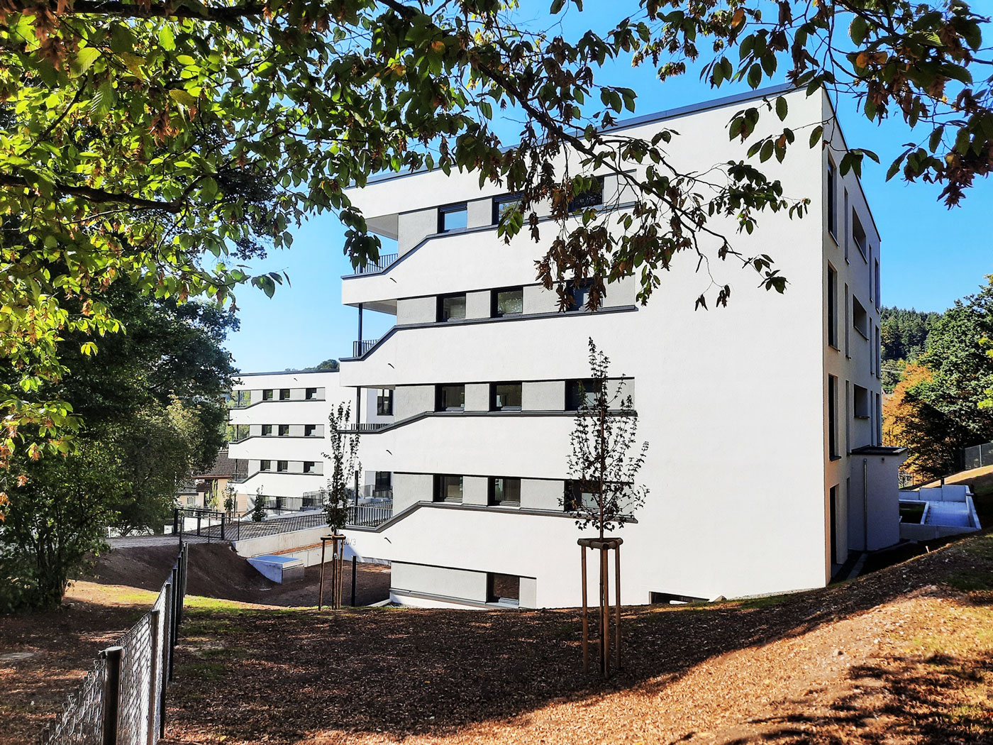 Schiesstal Schwäbisch Gmünd Ein modernes, weißes Mehrfamilienhaus von SCHATZ Wohnbau besticht durch kantige Balkone, umgeben von Bäumen und einer grasbewachsenen, abfallenden Fläche unter strahlend blauem Himmel – ideal für alle, die eine Wohnung kaufen möchten, oder für Investoren, die eine Immobilie erwerben möchten.