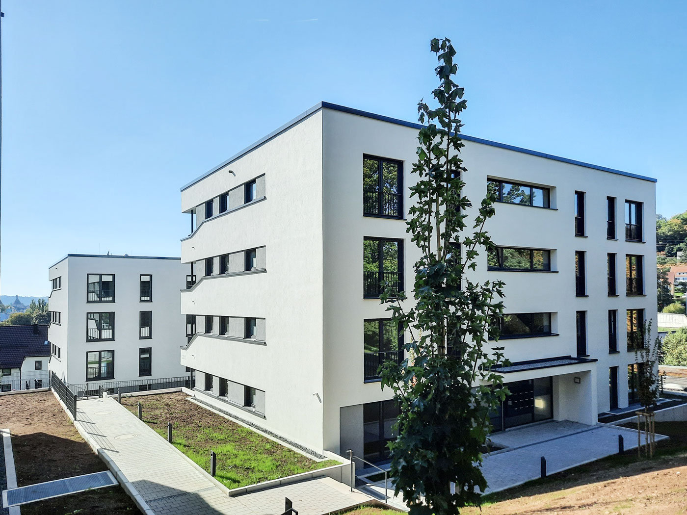 Schiesstal Schwäbisch Gmünd Unter dem strahlend blauen Himmel in Schorndorf stehen zwei moderne, weiße Neubau-Wohnhäuser mit großen Fenstern nebeneinander. Im Vordergrund sind ein junger Baum und gepflasterte Wege zu sehen, die die durchdachte Planung des Bauträgers widerspiegeln.
