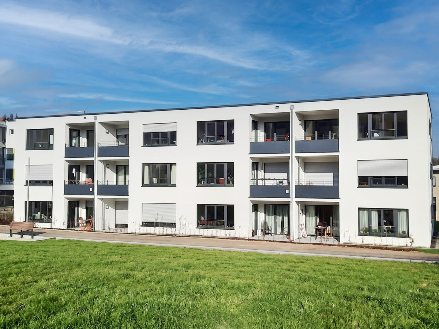 Betreutes Wohnen Schafhofstraße Albershausen Ein modernes, weißes, dreistöckiges Mehrfamilienhaus mit zahlreichen Balkonen und großen Fenstern, umgeben von grünem Rasen unter blauem Himmel – ein außergewöhnliches Projekt von SCHATZ Wohnbau, dem renommierten Bauträger mit Sitz in Schorndorf.