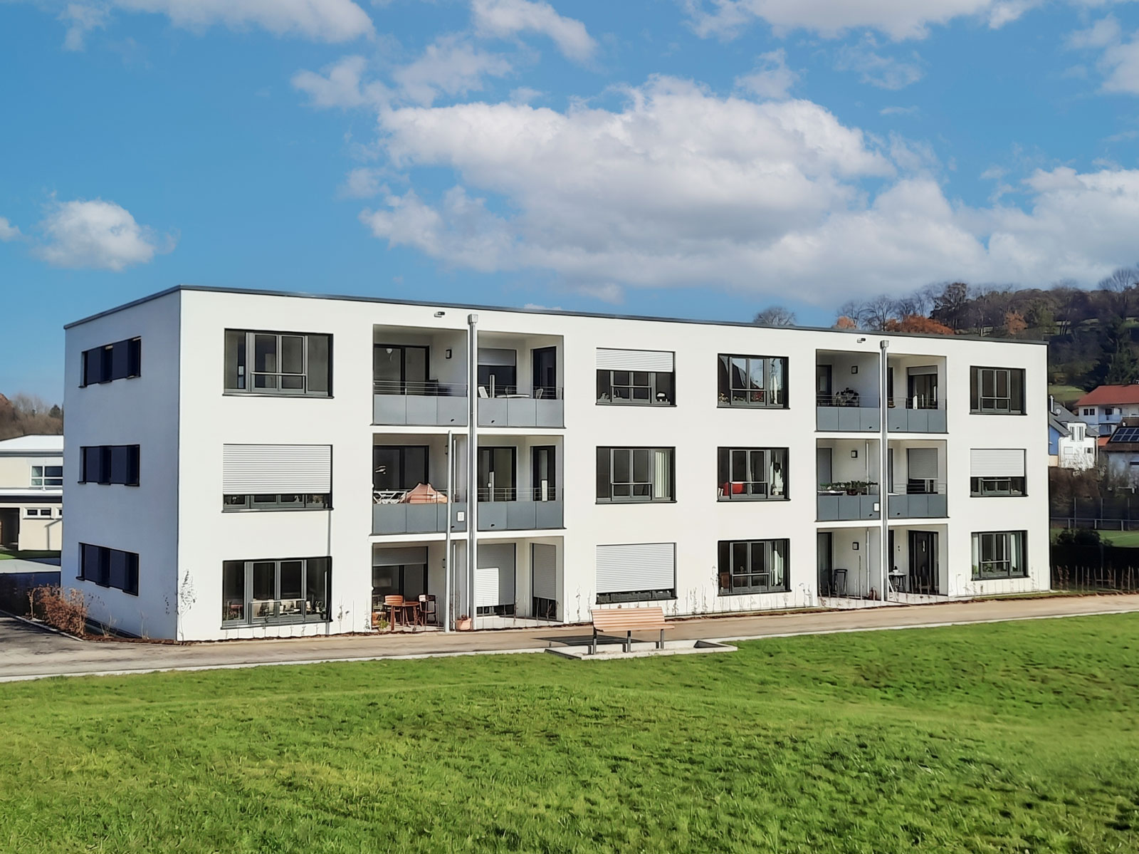 Ein modernes, dreistöckiges, weißes Wohnhaus von SCHATZ Wohnbau mit Balkonen und großen Fenstern hebt sich anmutig vor dem Hintergrund eines blauen, bewölkten Himmels ab. Das in Schorndorf gelegene Neubauobjekt verfügt über eine Rasenfläche und einen gepflasterten Weg vor dem Haus und verbindet so zeitgenössisches Design mit einladenden Außenbereichen.