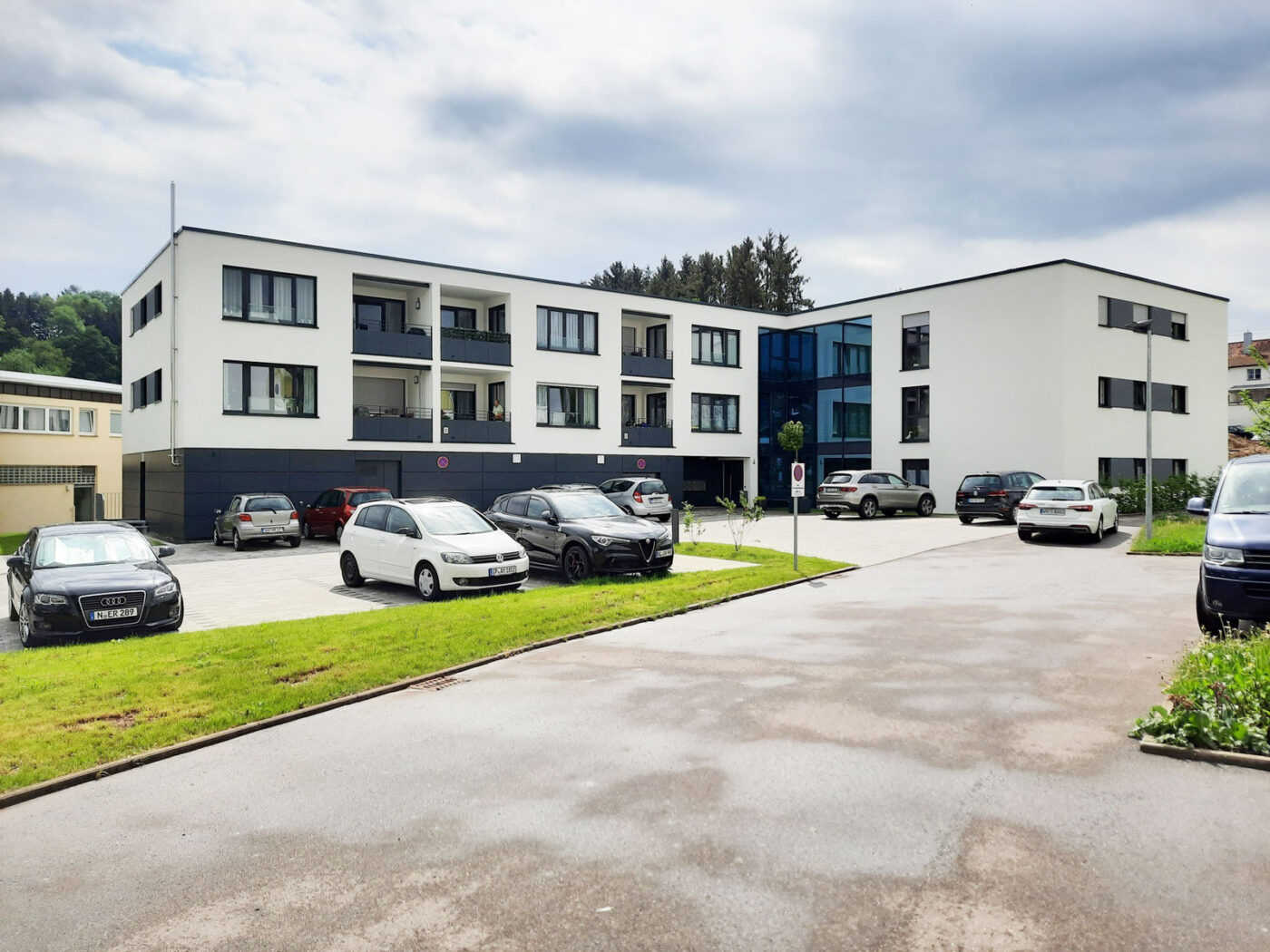 Betreutes Wohnen Schafhofstraße Albershausen Modernes, weißes, dreistöckiges Mehrfamilienhaus mit Balkonen und großen Fenstern, errichtet von SCHATZ Wohnbau. Vor dem Gebäude stehen mehrere Autos auf einem gepflasterten Parkplatz, während Bäume und Grünflächen das Haus unter einem teilweise bewölkten Himmel in Schorndorf umgeben.