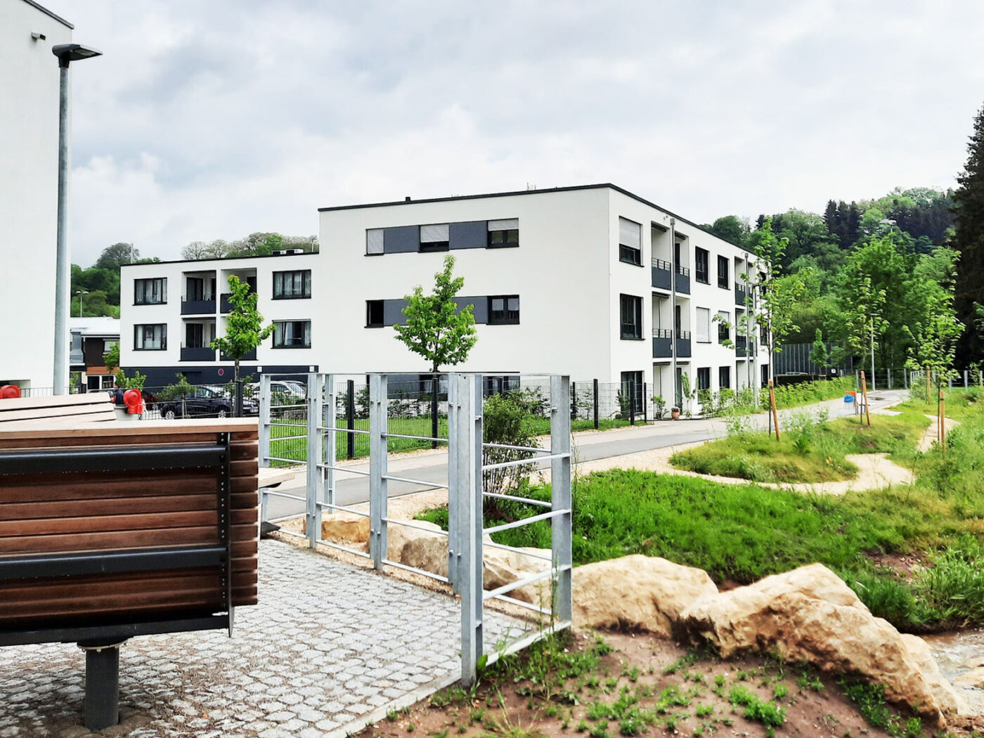 Betreutes Wohnen Schafhofstraße Albershausen Moderne weiße Wohnblocks eines renommierten Bauträgers stehen unter bewölktem Himmel in der Nähe einer landschaftlich gestalteten Grünfläche mit jungen Bäumen, einer Steinumrandung, einem Weg und einer Bank am Eingang – eine ideale Gelegenheit für alle, die in Schorndorf eine Immobilie kaufen möchten.