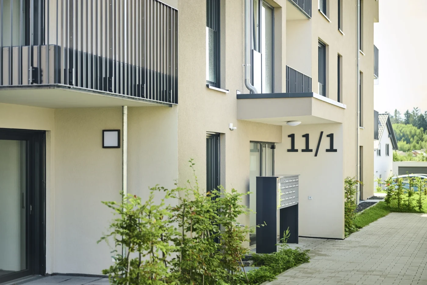 Modernes Mehrfamilienhaus mit beigen Wänden, Balkonen und der Hausnummer 11/1 am Eingang. Grüne Sträucher säumen den Gehweg, und neben der Tür sind Briefkästen zu sehen. Diese ruhige Wohnanlage, die von SCHATZ Wohnbau entwickelt wurde, bietet eine verlockende Gelegenheit für alle, die eine Wohnung von einem renommierten Bauträger kaufen möchten.