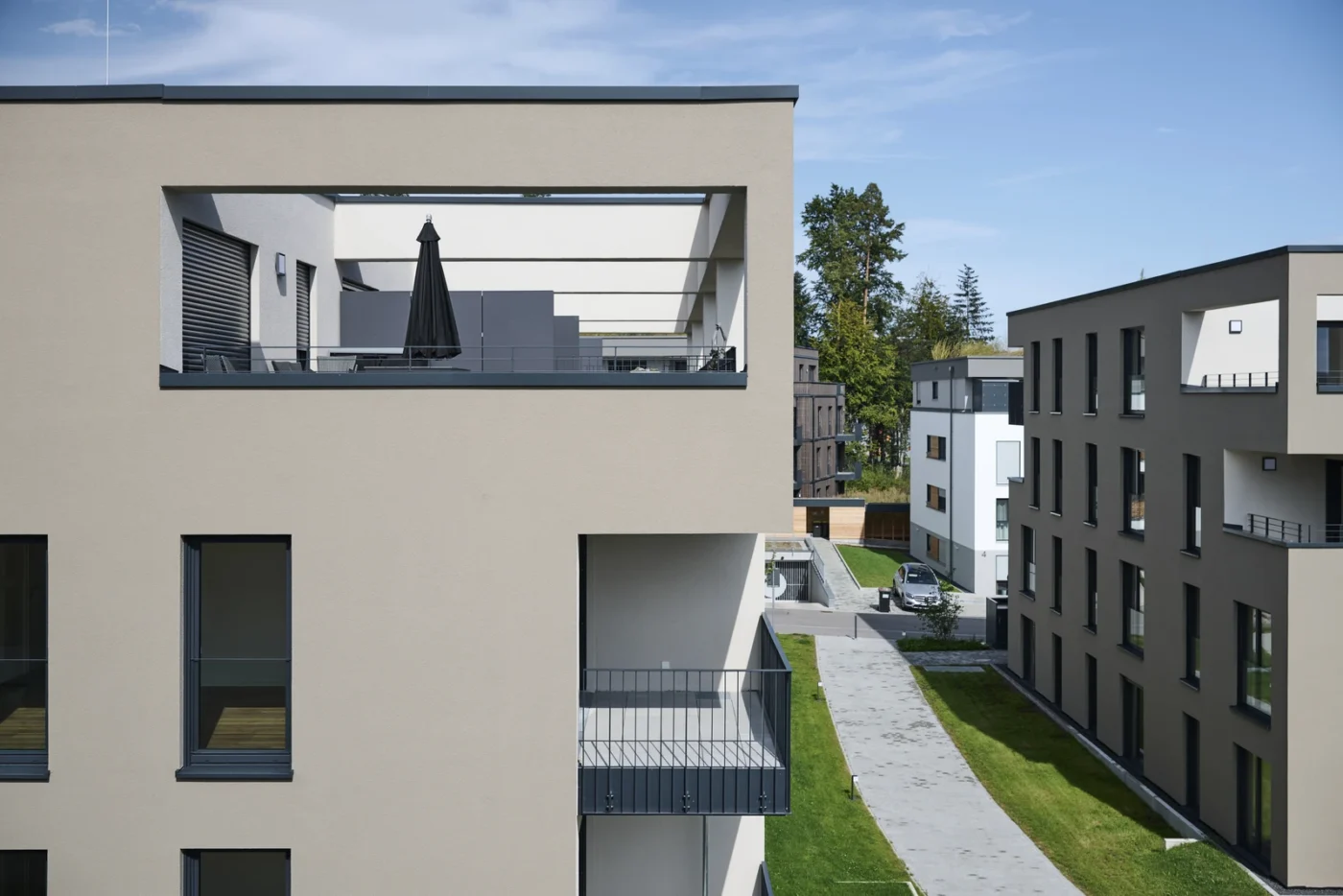Moderne Neubau-Wohnhäuser mit Balkonen und Terrassen, beigen Fassaden, großen Fenstern und einem Verbindungsgang dazwischen. Auf der oberen Terrasse sind ein schwarzer Sonnenschirm und Gartenmöbel zu sehen. Über den Häusern erstreckt sich ein strahlend blauer Himmel – ideal für alle, die eine Wohnung in einem zeitgemäßen Umfeld kaufen möchten.
