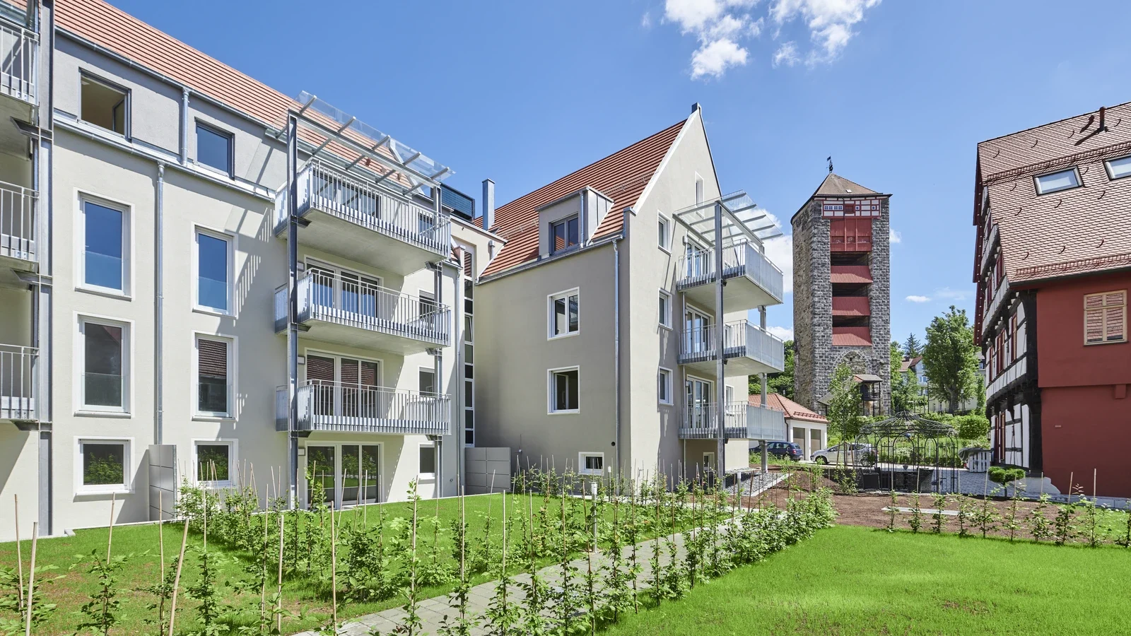 Leistungen Moderne Mehrfamilienhäuser mit Balkonen, roten Ziegeldächern und einem gepflegten grünen Rasen prägen diesen Neubau in Schorndorf. Im Hintergrund ragt ein alter Steinturm vor einem strahlend blauen Himmel mit vereinzelten Wolken empor und schafft so eine harmonische Verbindung aus zeitgemäßem Wohnen und historischem Charme – ein Vorzeigeprojekt eines erfahrenen Bauträgers.