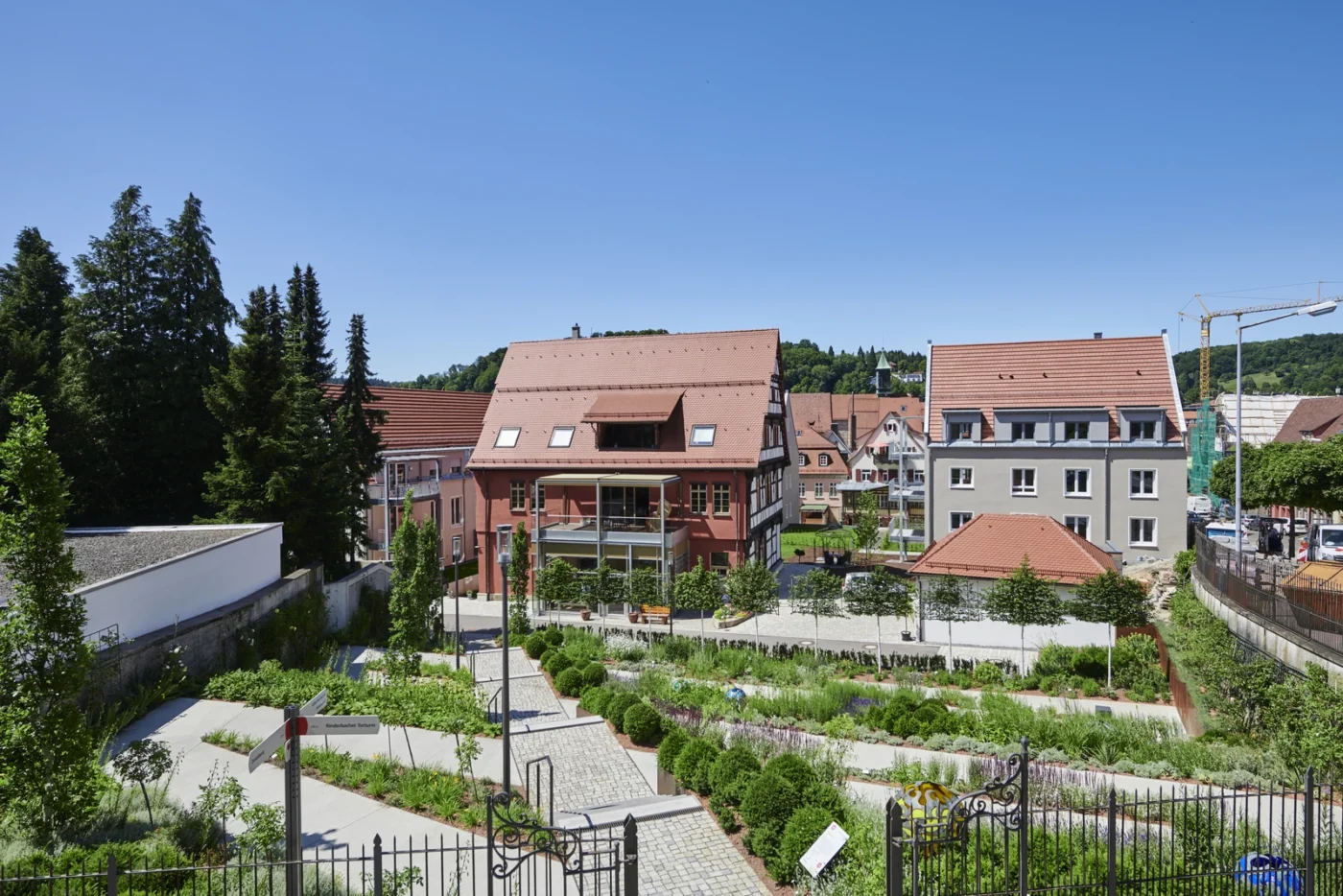 An einem sonnigen Tag erstreckt sich vor den rotgedeckten Neubauten ein gepflegter Gemeinschaftsgarten mit Wegen. Im Hintergrund sind unter einem strahlend blauen Himmel Bäume und weitere Gebäude zu sehen.