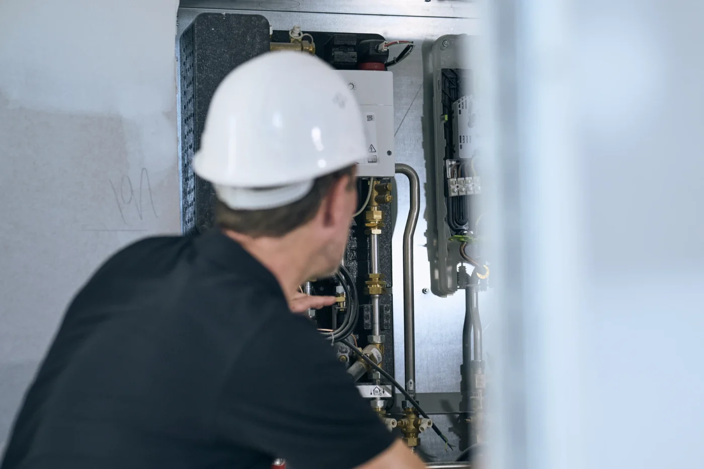 Qualitätssicherung Eine Person mit weißem Schutzhelm und schwarzem Hemd begutachtet Rohre und elektrische Komponenten in einem Industrieschaltkasten und zeigt darauf – ein Beispiel für die Liebe zum Detail, die bei den Neubau-Projekten von SCHATZ Wohnbau unverzichtbar ist.