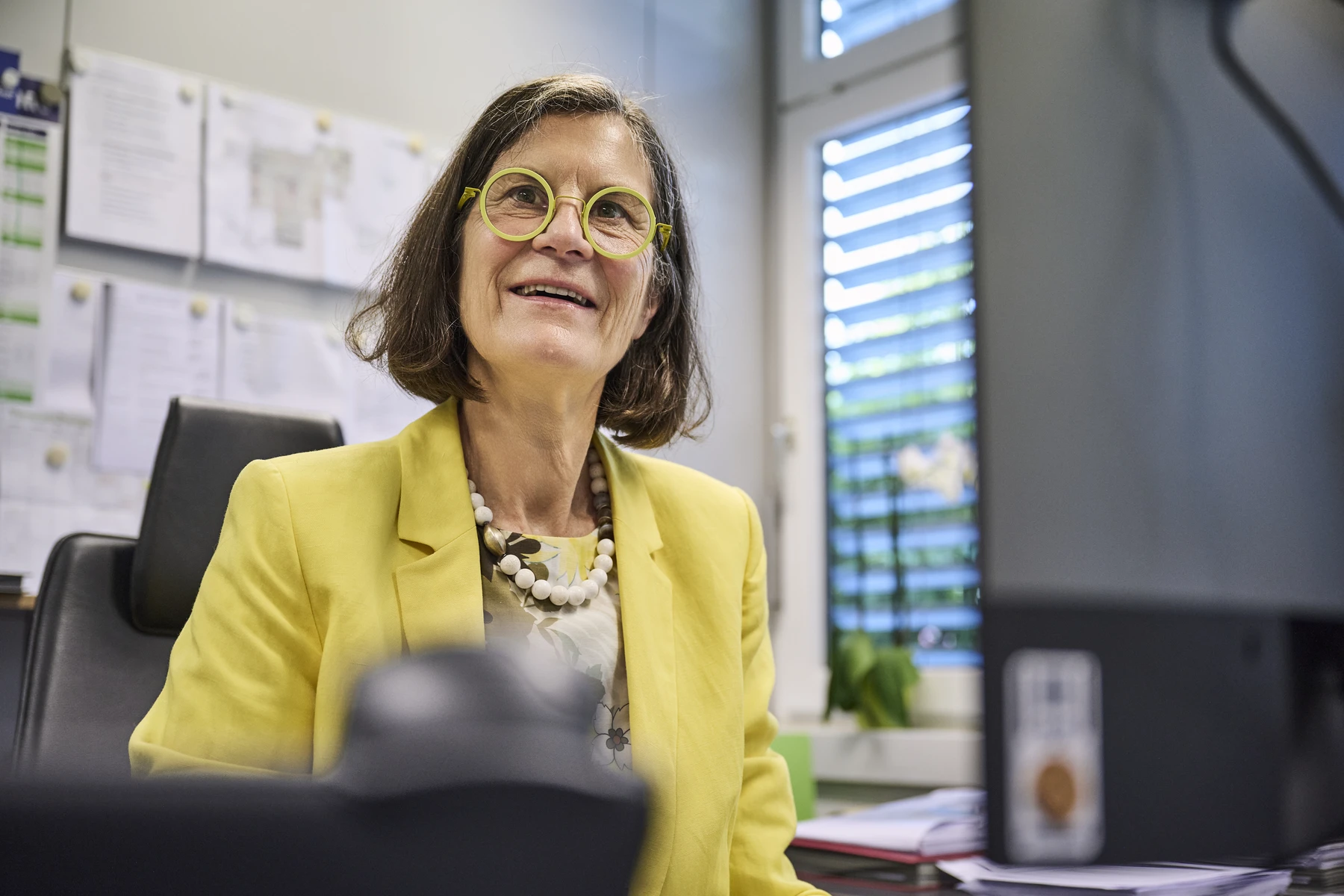 Immobilie als Kapitalanlage für Investoren Eine Frau mit schulterlangem braunem Haar, die eine runde Brille und einen gelben Blazer trägt, sitzt an einem Schreibtisch in einem Büro, in dem Papiere an der Wand hängen und hinter ihr ein Fenster mit Jalousien zu sehen ist – vielleicht prüft sie gerade Unterlagen für Kunden, die an einem Neubau interessiert sind oder eine Wohnung in Schorndorf kaufen möchten.