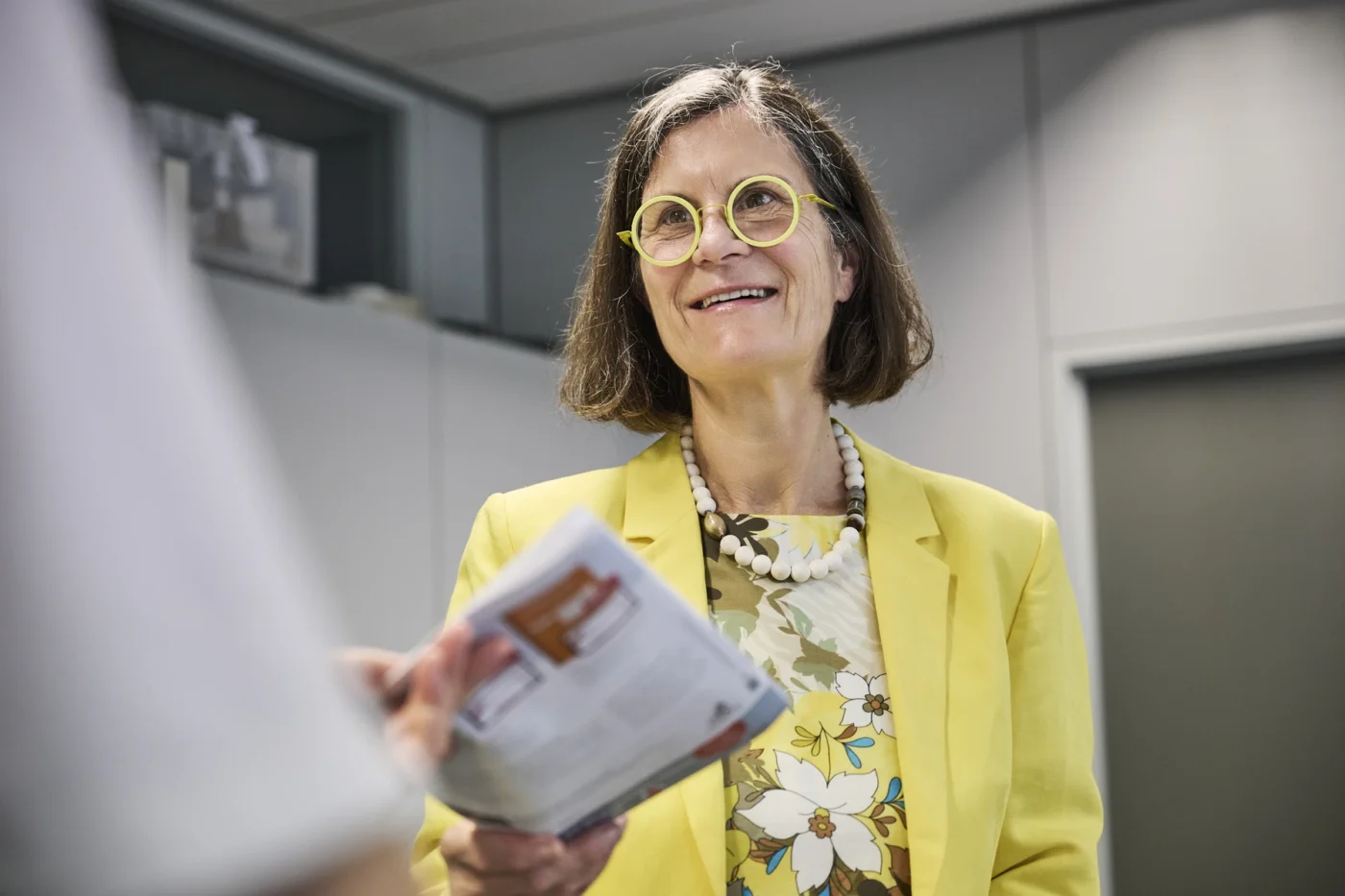 Vorteile Eine Frau mit gelber Brille und gelbem Blazer lächelt, während sie in einem Büro eine Zeitschrift oder Broschüre in der Hand hält; möglicherweise bespricht sie gerade mit einem Bauträger die Möglichkeiten, eine Immobilie in Schorndorf zu kaufen.
