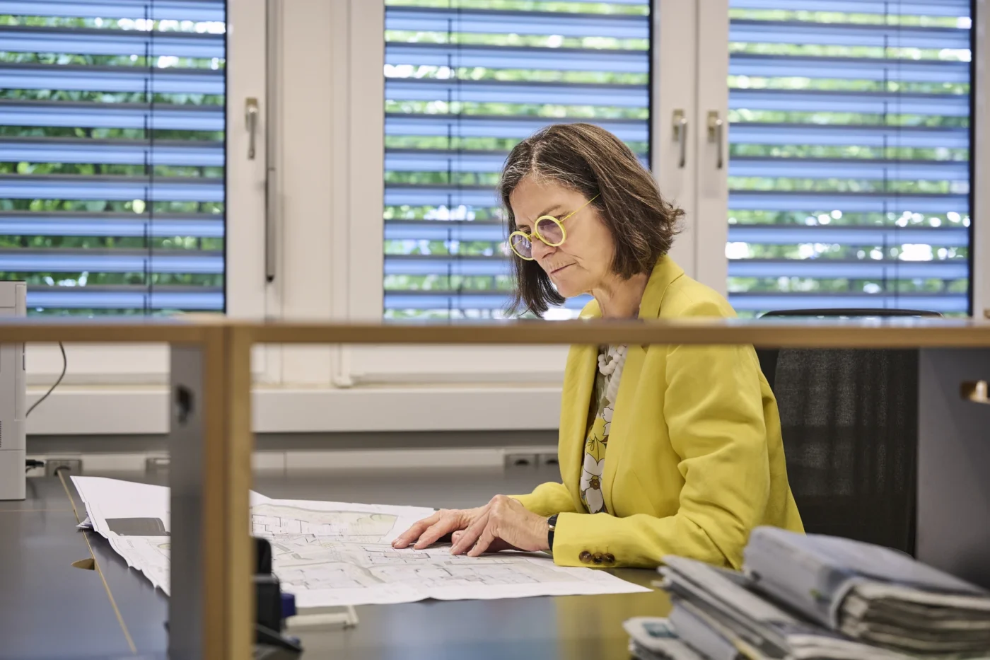 Immobilie als Kapitalanlage für Investoren Eine Frau in einem gelben Blazer und mit Brille sitzt an einem Schreibtisch in einem Büro und prüft Baupläne für SCHATZ Wohnbau, einen renommierten Bauträger mit Sitz in Schorndorf. Hinter ihr strömt Tageslicht durch große Fenster mit Jalousien herein.