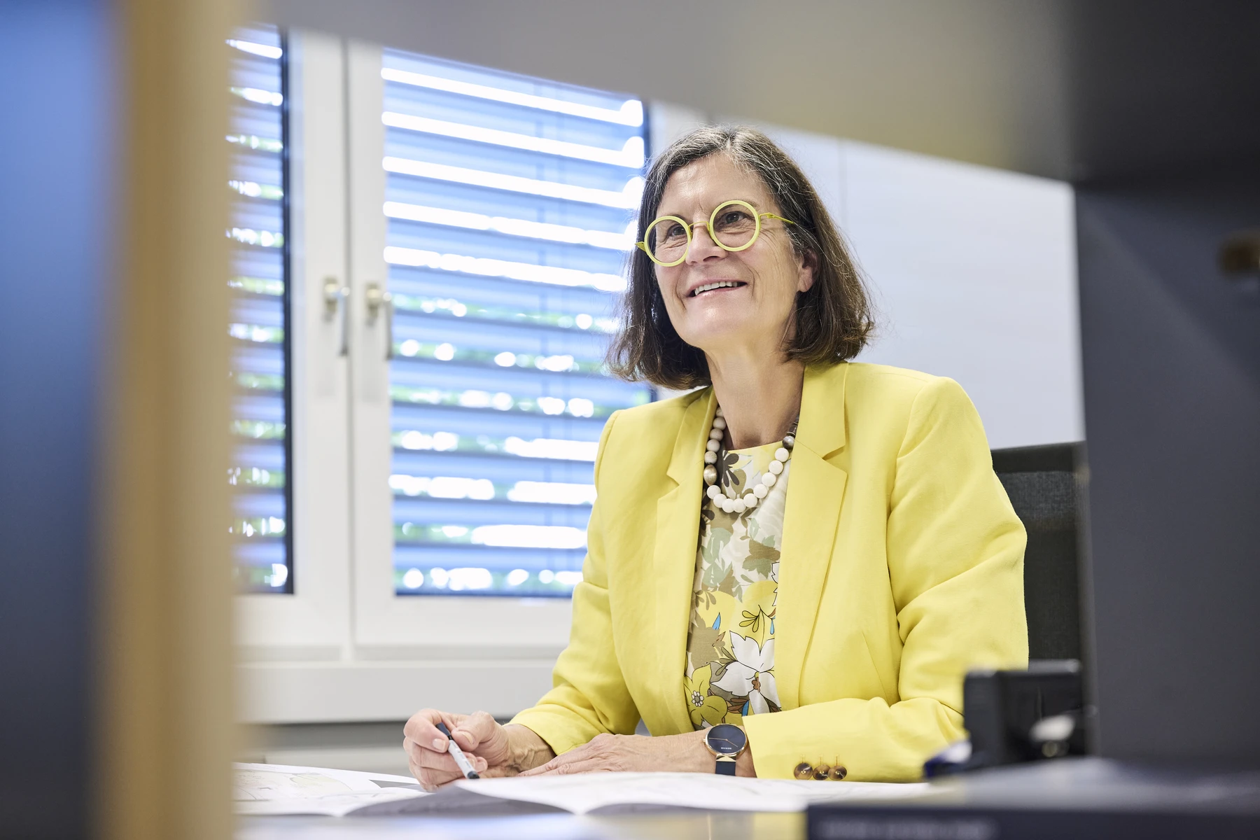 Leistungen Eine Frau in einem gelben Blazer und mit Brille sitzt an einem Schreibtisch und lächelt, während sie einen Stift in der Hand hält. Sie befindet sich in einem hellen Büro mit großen Fenstern und Fensterläden im Hintergrund und bespricht möglicherweise Neubauvorhaben mit Kunden, die sich für den dynamischen Bauträgermarkt in Schorndorf interessieren.