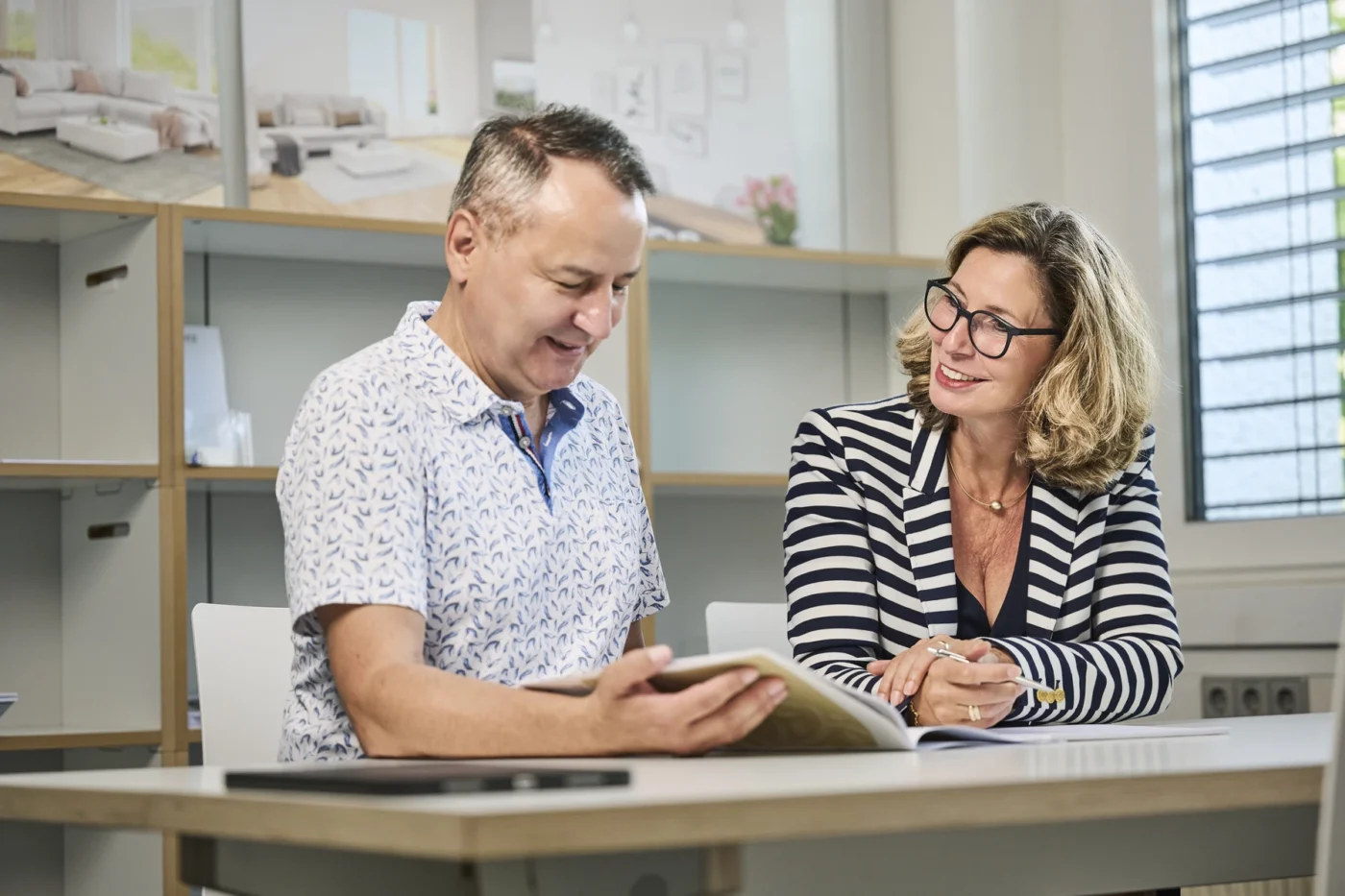 Ausstattung & Planung Zwei Erwachsene sitzen an einem Tisch in einem hellen Büro in Schorndorf und schauen sich gemeinsam ein Notizbuch an. Der Mann lächelt beim Lesen, und die Frau von SCHATZ Wohnbau, die eine Brille trägt, lächelt ihn an. Im Hintergrund sind Regale und Fotos zu sehen.