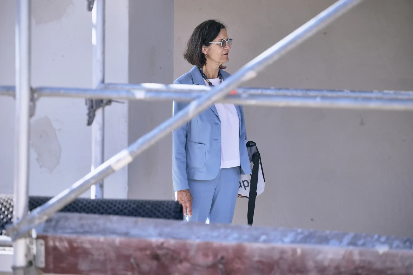 Leistungen Eine Frau in einem blauen Kostüm und mit Sonnenbrille steht neben einer Wand, die teilweise von einem Gerüst verdeckt wird – vielleicht bei einem Neubauprojekt von SCHATZ Wohnbau in Schorndorf – und hält eine Tasche und einen Regenschirm in der Hand.