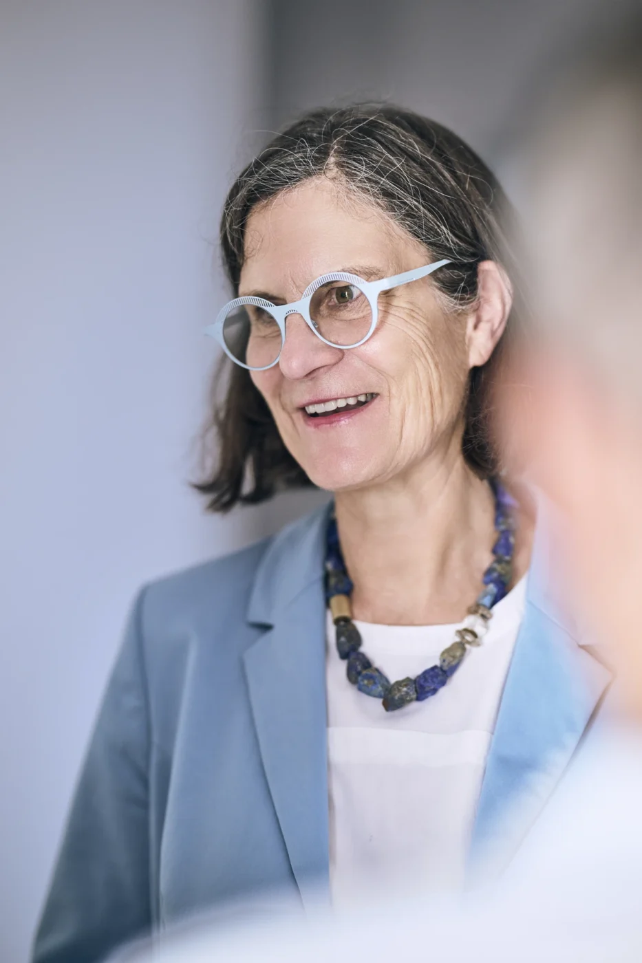 Ausstattung & Planung Eine Frau mit schulterlangem braunem Haar, die eine blaue Brille, einen blauen Blazer, ein weißes Oberteil und eine klobige Perlenkette trägt, lächelt, während sie mit jemandem außerhalb des Bildausschnitts spricht – vielleicht besprechen sie gerade die spannende Gelegenheit, eine Immobilie zu kaufen, oder tauschen sich über Neubau-Projekte aus.