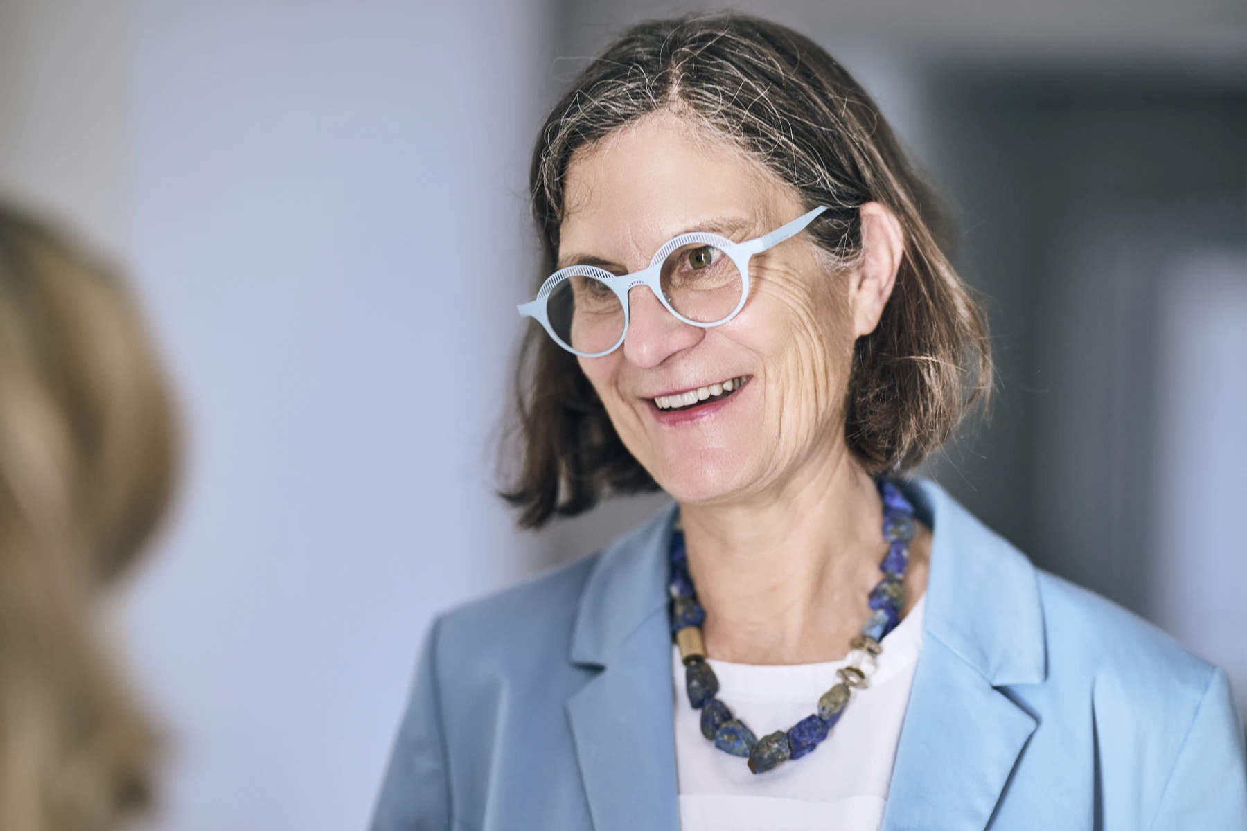 Energieeffiziente Neubauwohnung kaufen Eine Frau mit schulterlangem braunem Haar, die eine hellblaue Brille, einen dazu passenden blauen Blazer und eine klobige blaue Halskette trägt, lächelt; die Aufnahme zeigt sie in einem Innenraum mit unscharfem Hintergrund, im Vordergrund ist eine weitere Person zu sehen – vielleicht besprechen sie gerade Möglichkeiten, eine Immobilie zu kaufen oder sich eine Neubauwohnung zu kaufen.