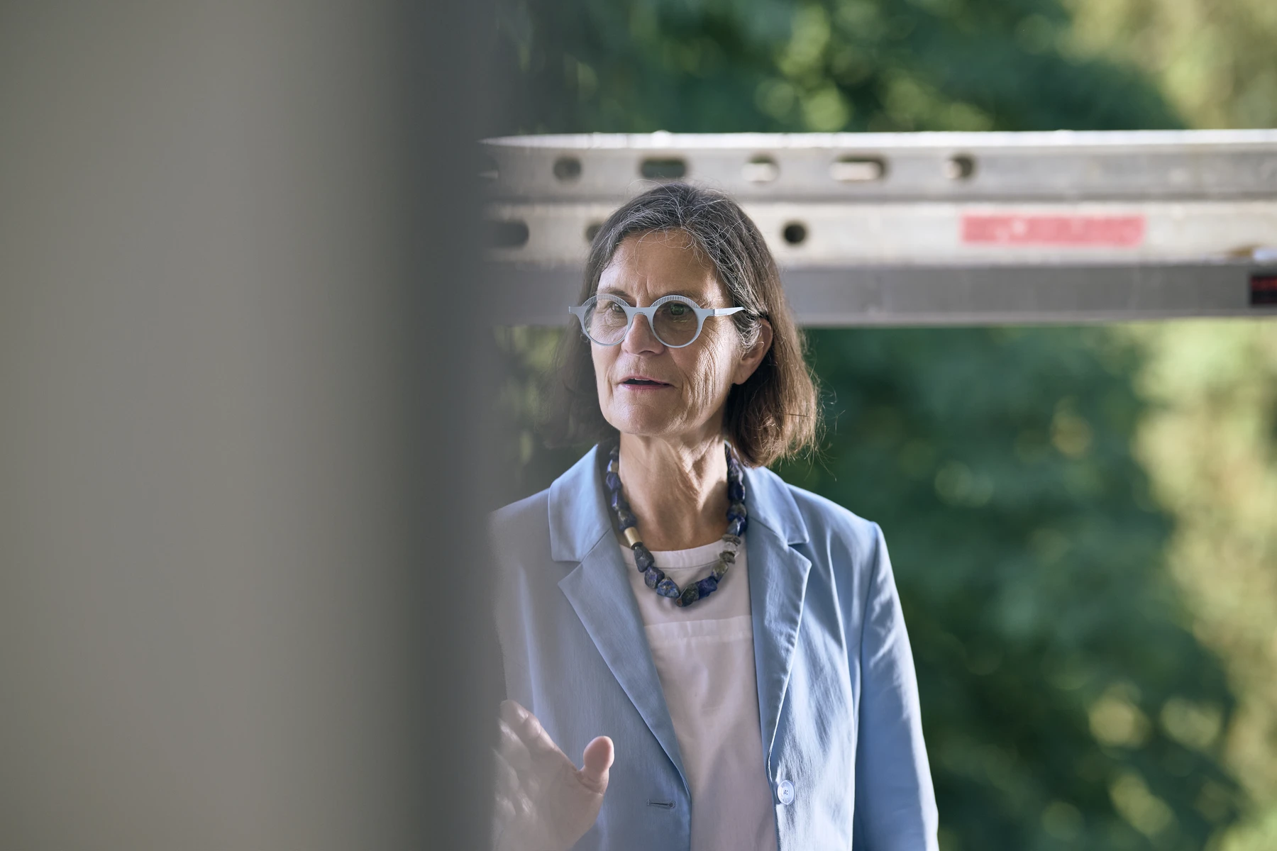 Immobilie als Kapitalanlage für Investoren Eine Frau mit schulterlangem braunem Haar, Brille und einem blauen Blazer steht im Freien und unterhält sich möglicherweise über SCHATZ Wohnbau oder darüber, wie man eine Neubauimmobilie kauft. Im Hintergrund ist Grün zu sehen, im Vordergrund ein unscharfes, vertikales Objekt. Sie scheint zu sprechen oder zu gestikulieren.