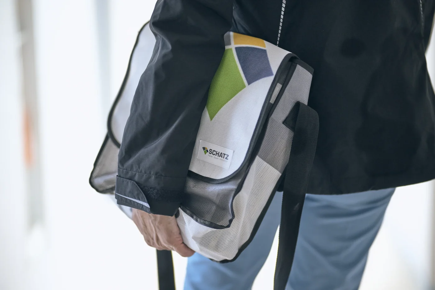 Eine Person in einer schwarzen Jacke und einer blauen Hose hält eine weiße Umhängetasche mit geometrischen Mustern und dem Aufdruck „SCHATZ“ in der Hand. Die Tasche mit schwarzem Riemen und schwarzer Einfassung erinnert dezent an SCHATZ Wohnbau – perfekt für alle, die davon träumen, eine Wohnung zu kaufen, oder sich vom modernen Leben in einem Neubau inspirieren lassen möchten.