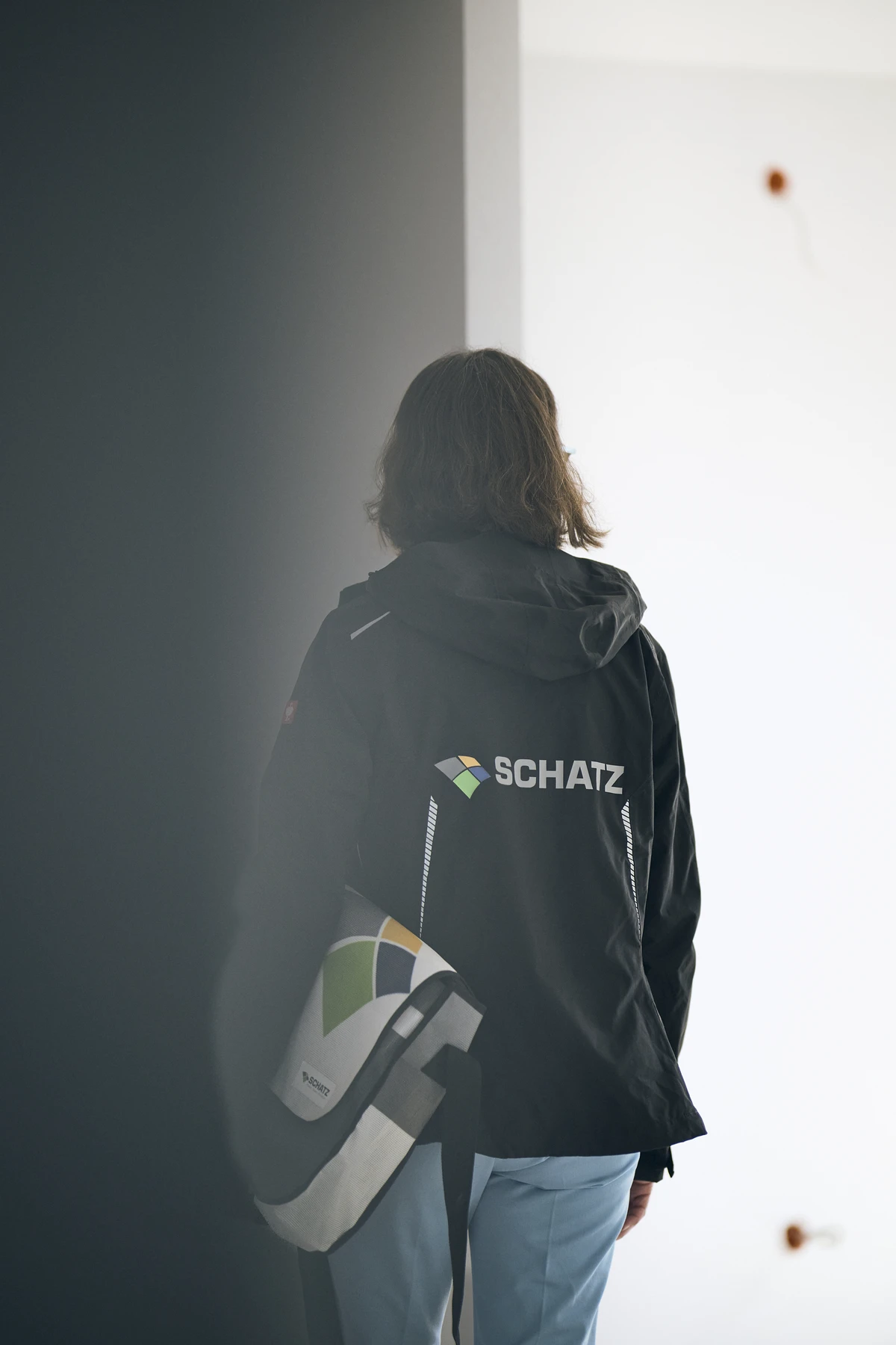 Über uns Eine Person mit schulterlangem Haar, die eine schwarze Jacke mit der Aufschrift „SCHATZ Wohnbau“ und einem geometrischen Logo auf dem Rücken trägt, steht in einem Innenraum und hält eine dazu passende schwarz-weiße Tasche in der Hand. Die Szene wirkt modern und minimalistisch und spiegelt die klare Ästhetik wider, die oft mit Neubau-Projekten in Verbindung gebracht wird.