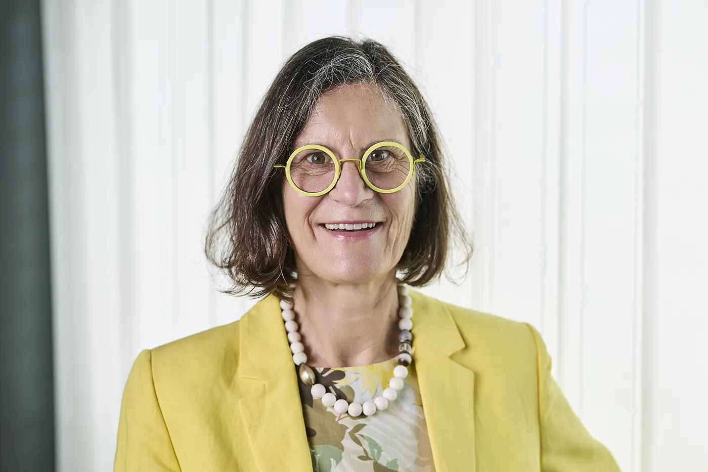Löwenseestraße Eine Frau mit schulterlangem braunem Haar, einer gelben Brille und einem gelben Blazer lächelt in die Kamera. Sie trägt ein geblümtes Oberteil und eine klobige weiße Perlenkette. Im Hintergrund sind weiße Vorhänge zu sehen.